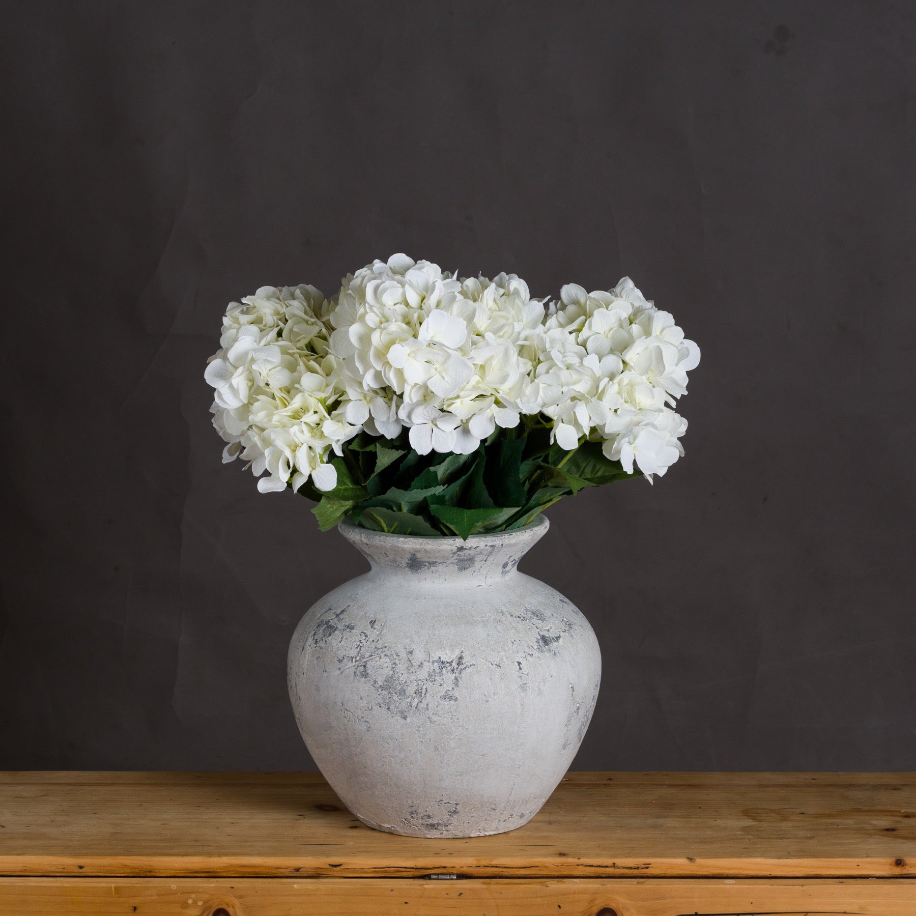 White hydrangeas in a textured vase on a wooden surface with a dark background