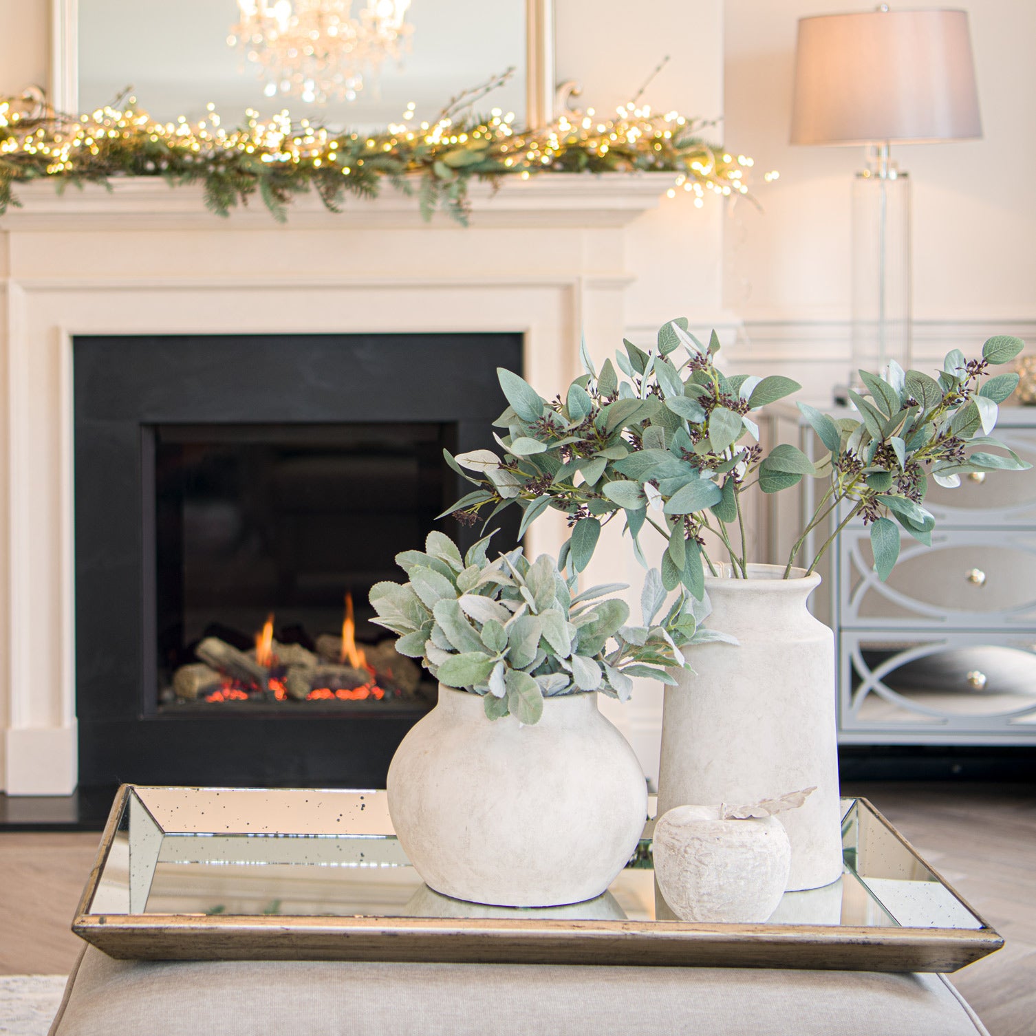 Decorative arrangement of vases with greenery on a coffee table in front of a fireplace.