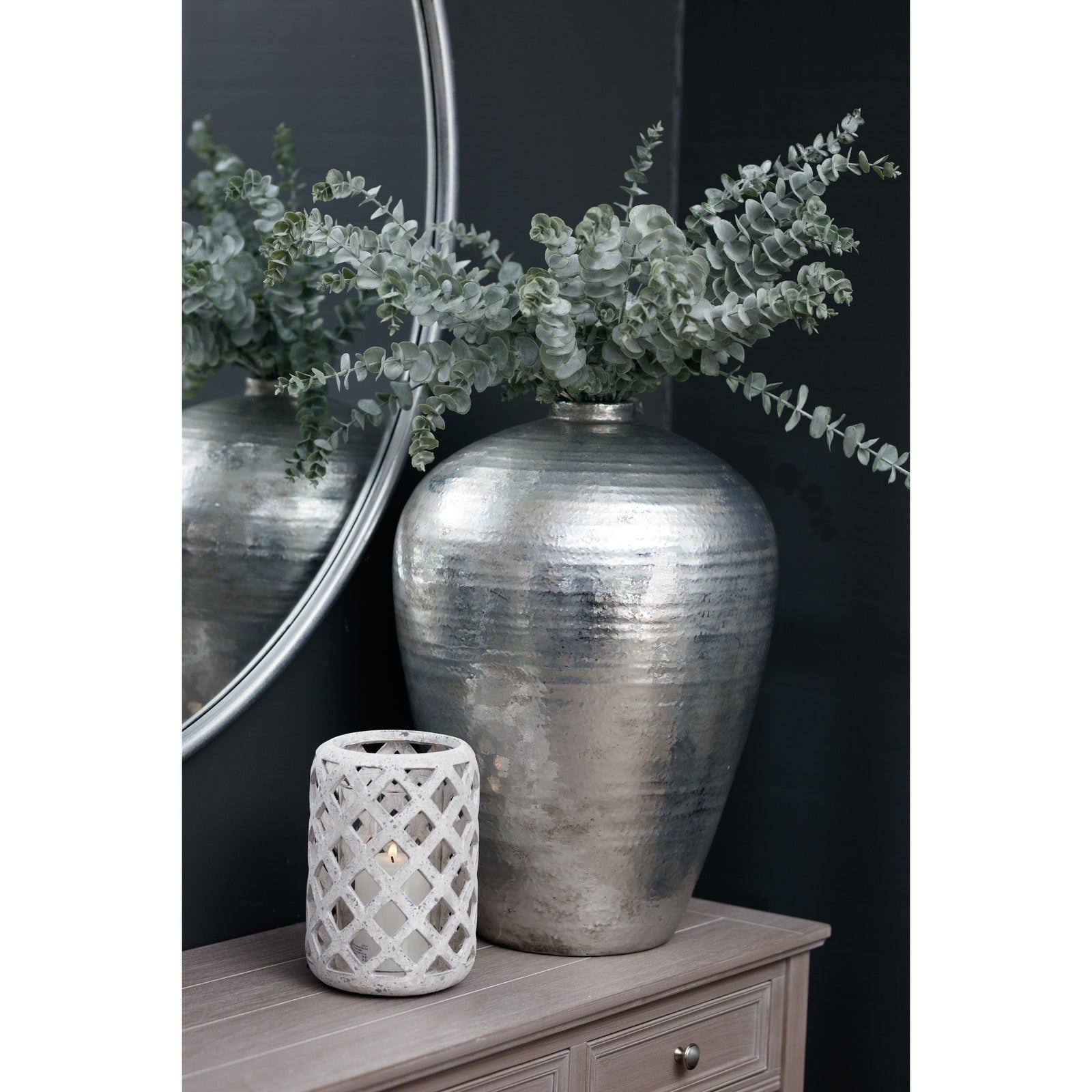 Silver textured vase with eucalyptus branches on a wooden surface against a dark wall.