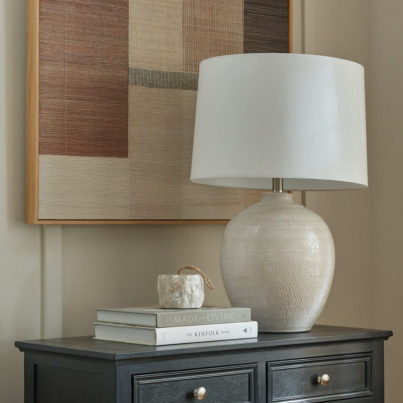 Beige textured lamp on a dark wooden side table with books and a small decorative item, against a neutral wall with a framed abstract art piece.
