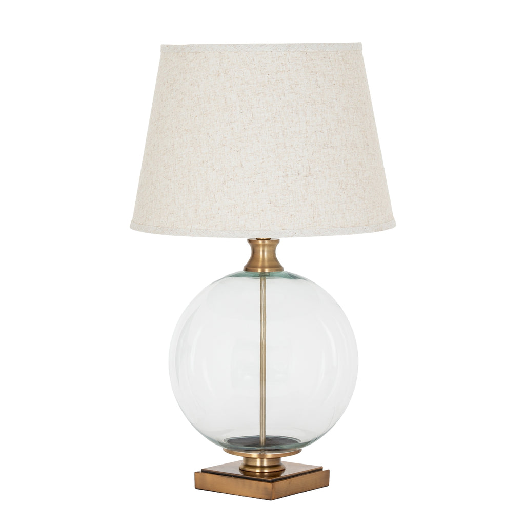 Table lamp with a clear glass base, brass accents, and a beige lampshade on a white background